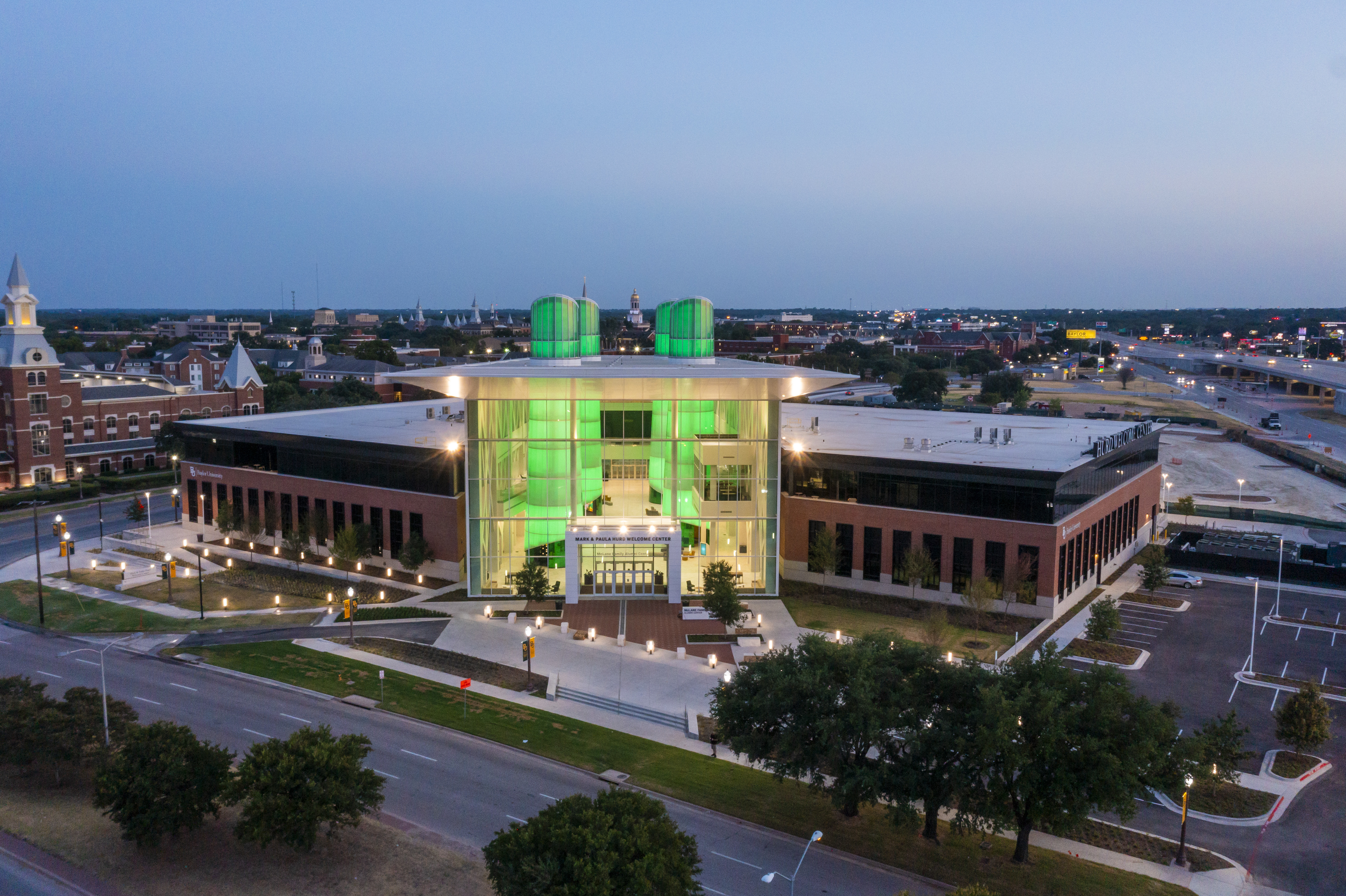 Arial view of the Hurd Welcome Center at night. The building's main towers are glowing in Baylor green lights. 