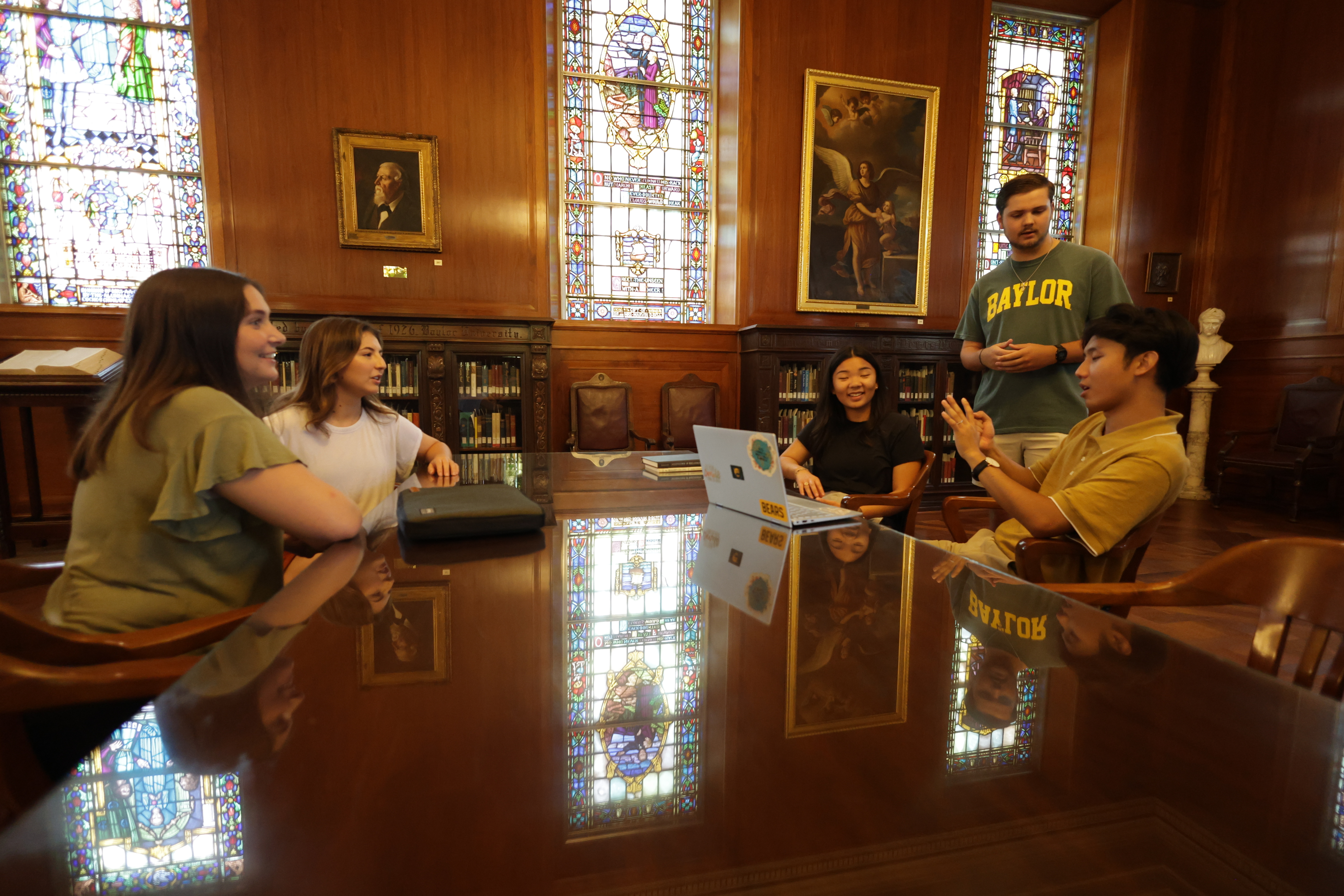 5 smiling students sit at a table with laptops open in the Armstrong Browning Library. The room is decorated with stained glass and dark woods.