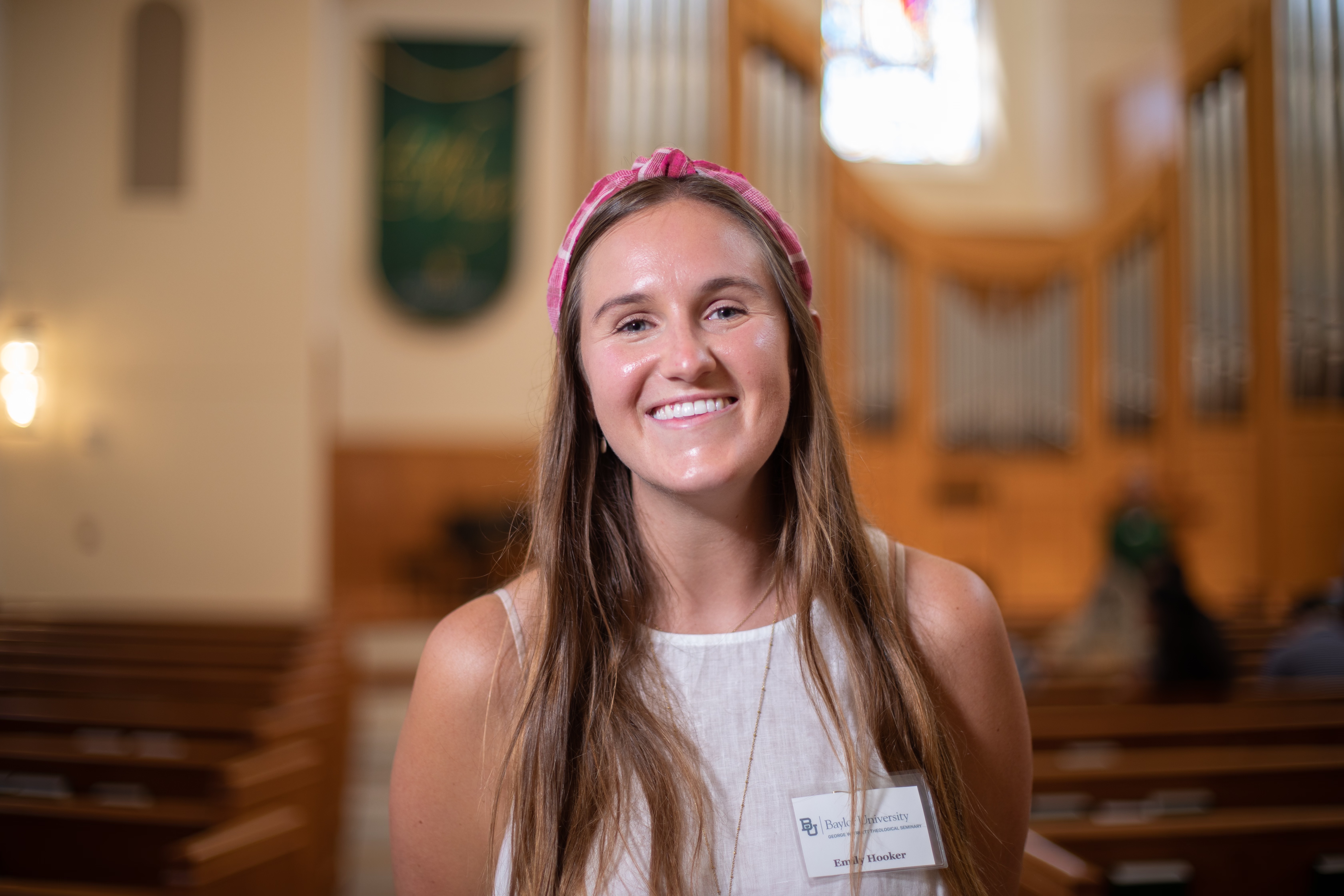 Emily Hooker stands in a white tank top and pink headband in the Truett Chapel. The background is blurred and Emily is seen smiling from mid chest up. 
