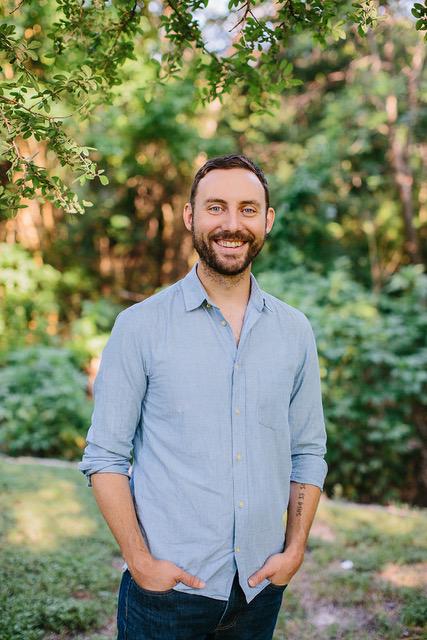 Sam Davidson stands outside in a field, surrounded by sunlight and trees. Photographed from the waist above, Davidson faces the camera, smiling in a blue collared shirt with his hands in his dark jeans.