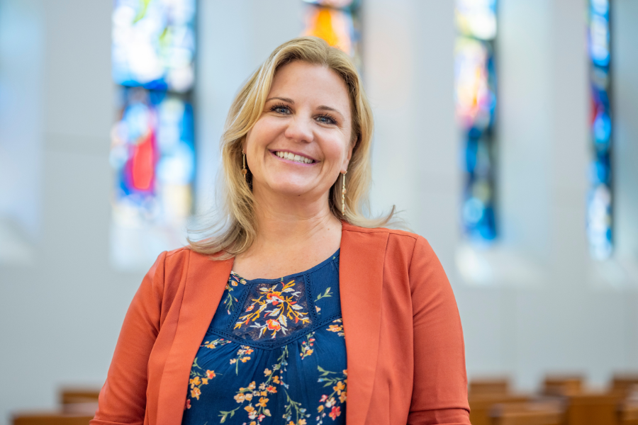 Dr. Jenny Matheny stands smiling at the camera. She is seen from mid torso up, wearing a orange jacket and a navy, floral blouse. Behind her, in blurred focus, are rows of pews and stained glass.